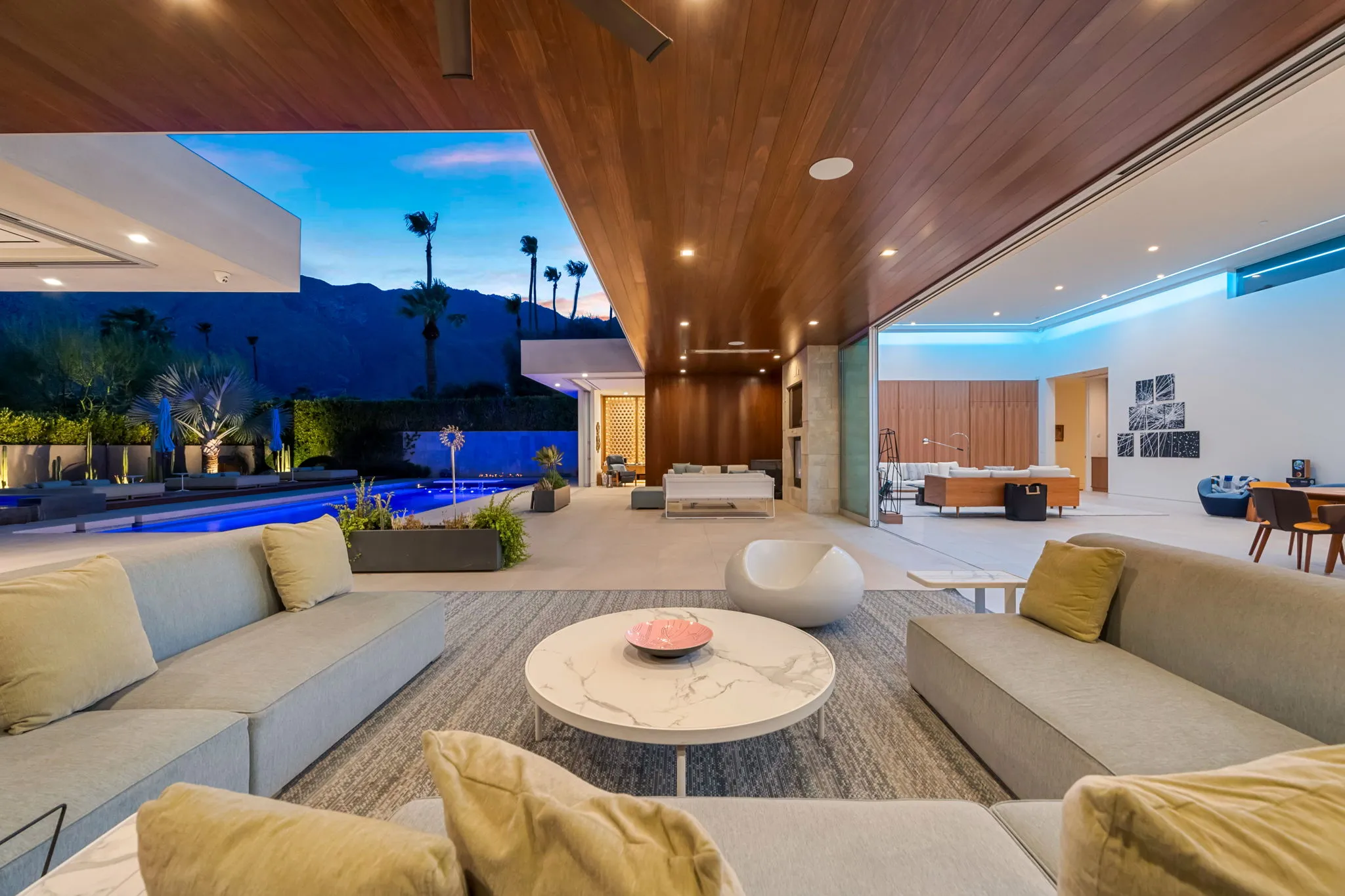 Covered patio at dusk with a wood-clad ceiling, lounge seating and mountain views beyond the pool
